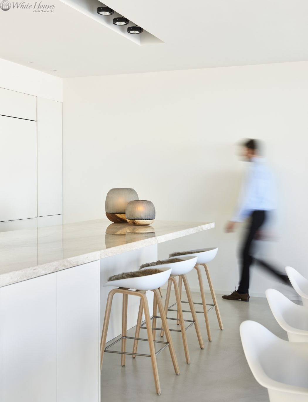 A modern, minimalist kitchen interior with sleek furniture, recessed lighting, and a blurred figure.