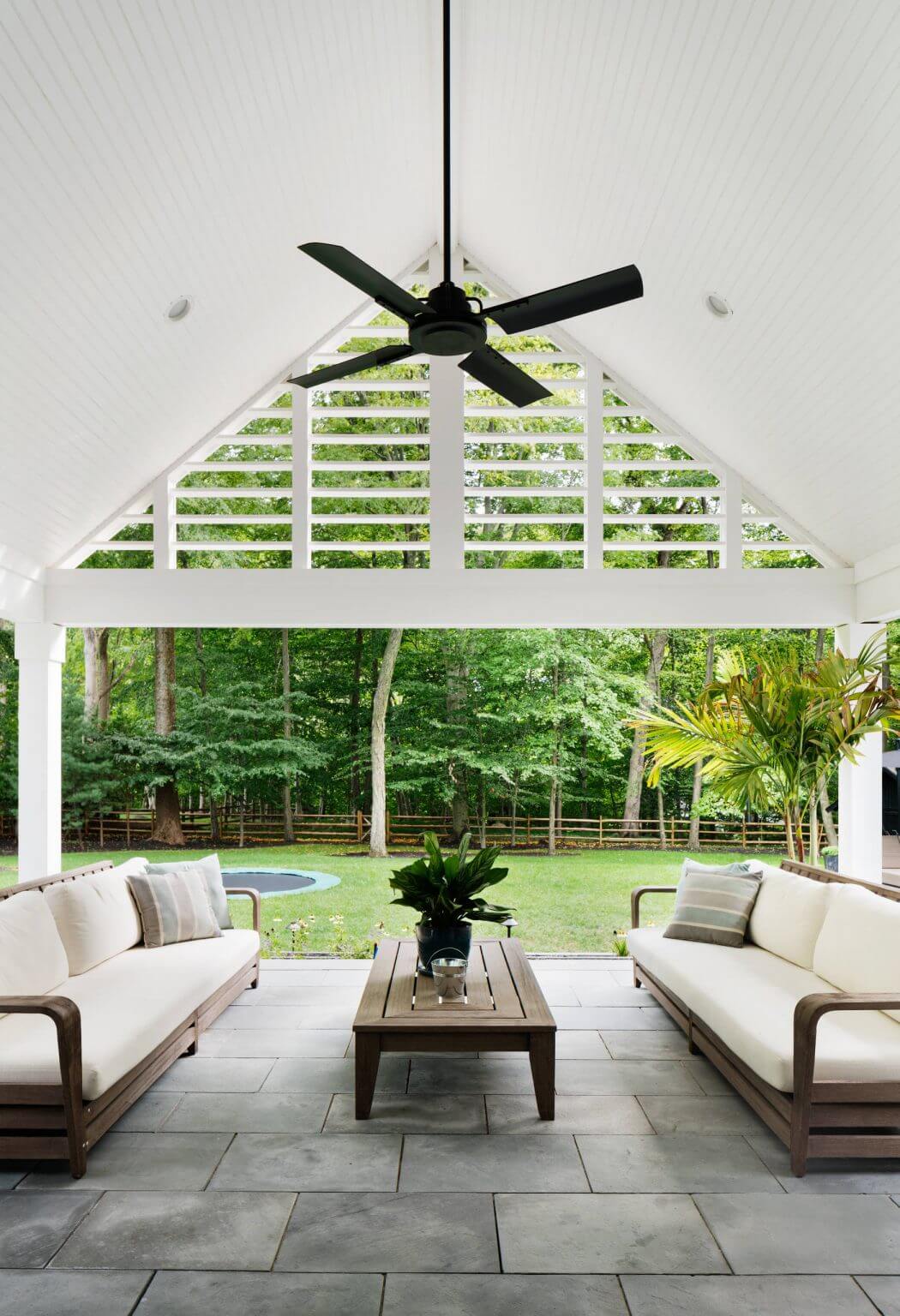 A modern outdoor patio with a vaulted ceiling, ceiling fan, and lush greenery surrounding it.