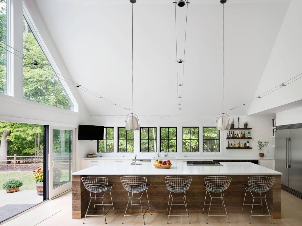 A bright, modern kitchen with large windows, sleek furnishings, and a wooden island with wire chairs.
