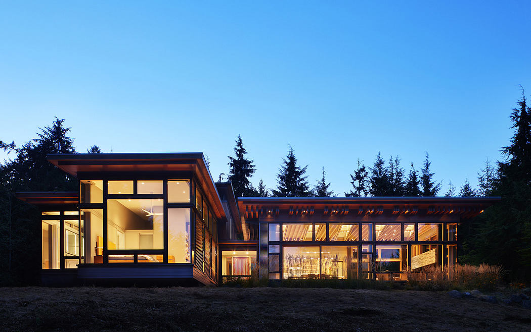 Modern house with large windows lit up at dusk against a forest backdrop.