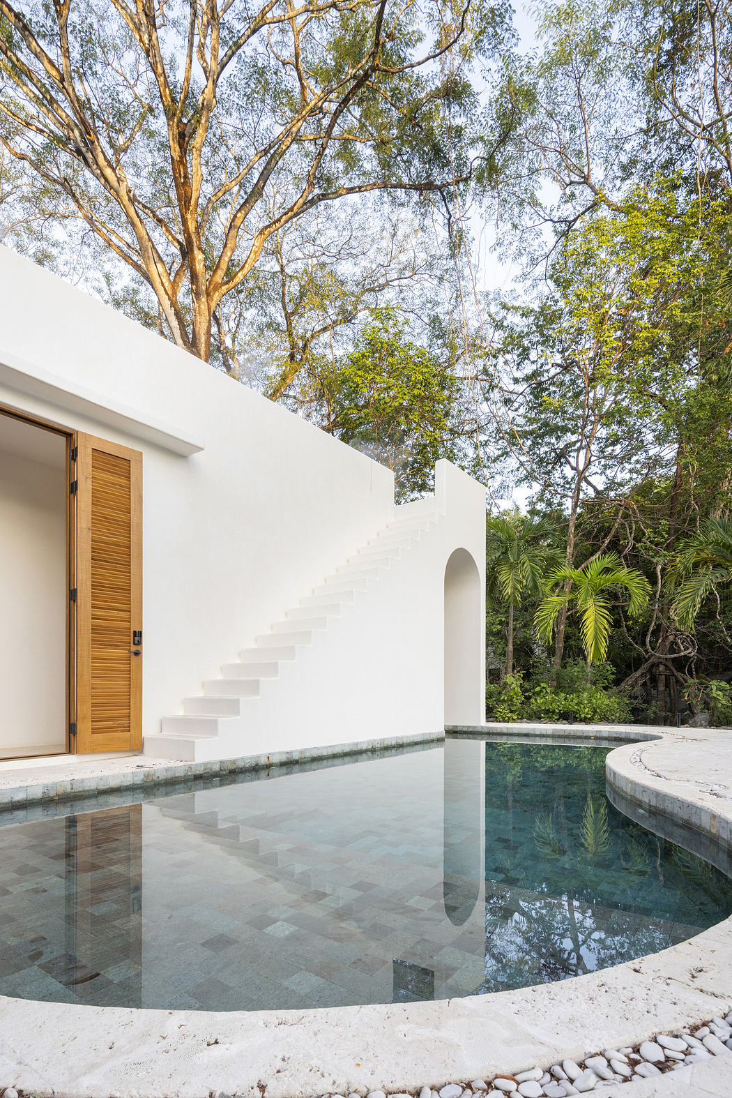 White modern house with an external staircase beside a circular pool, surrounded by trees.