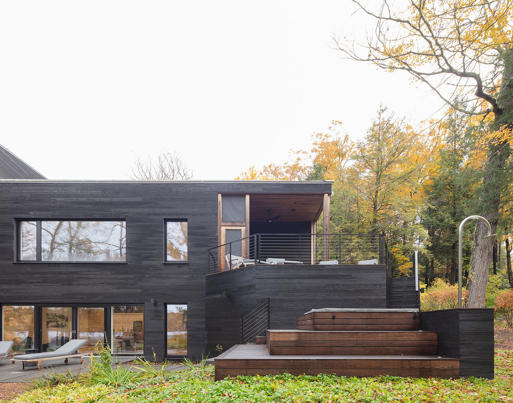 Modern black wooden house with large windows and outdoor deck surrounded by autumn trees.