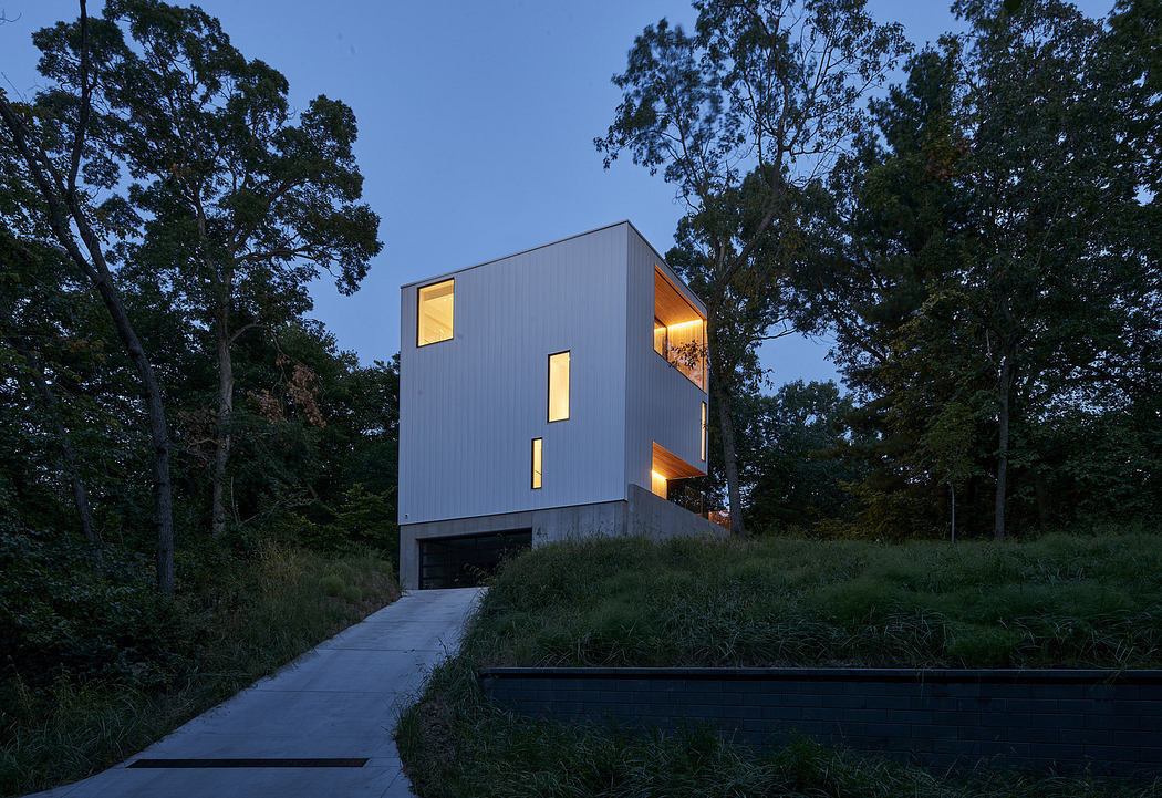 Modern minimalist house with illuminated windows at dusk surrounded by trees.