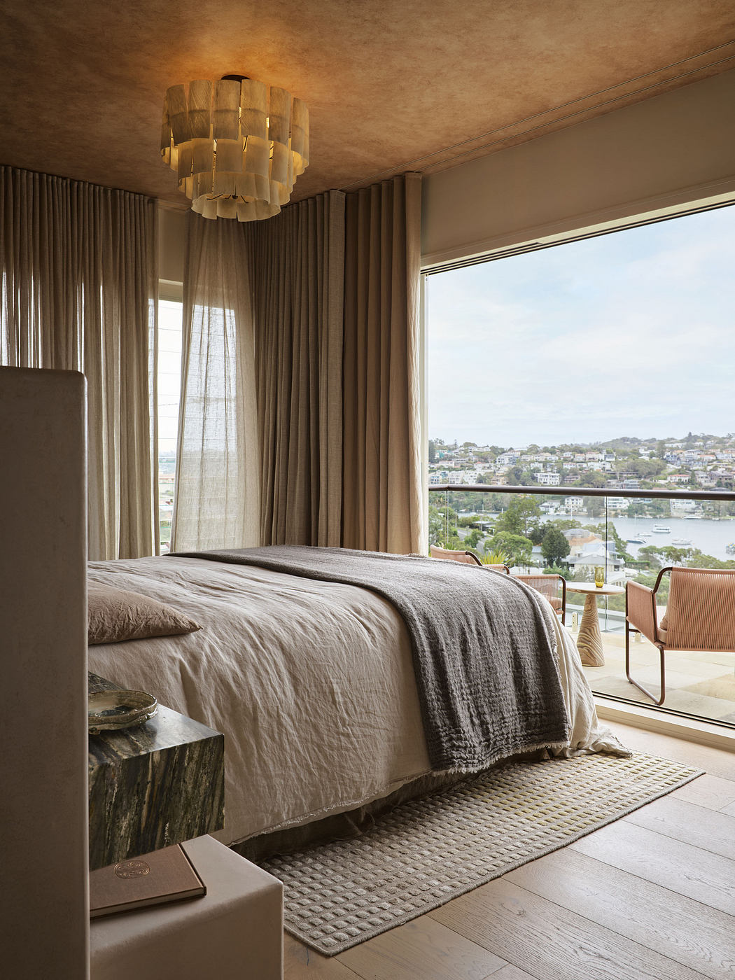 Elegant bedroom with large window overlooking water, neutral tones, and modern lighting.