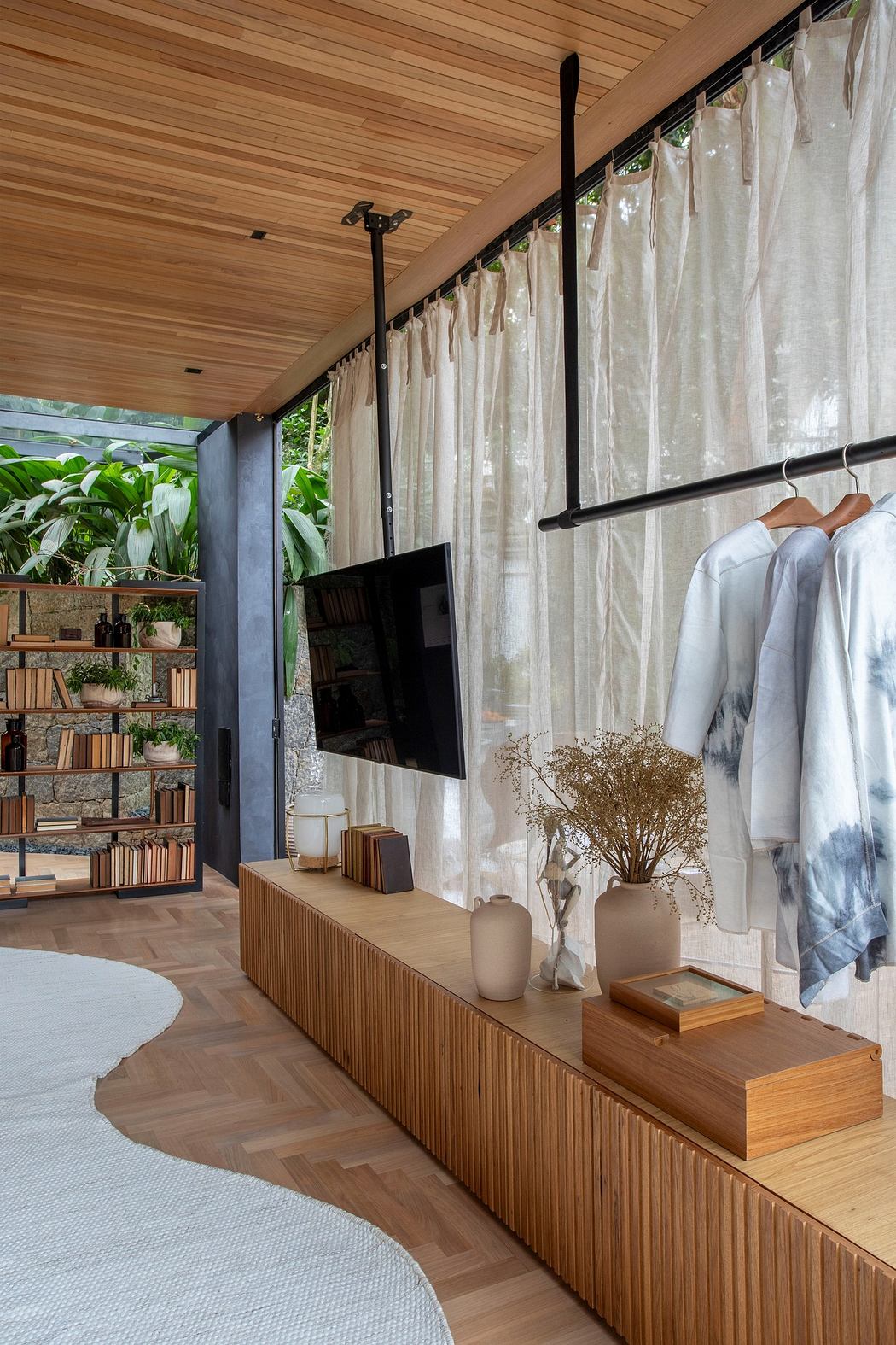 Modern room with wooden finish, bookshelf, hanging clothes, and indoor plants.