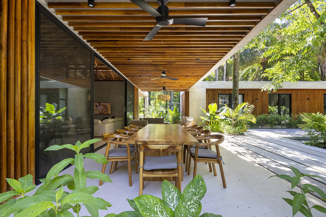 Open-air dining area with wooden table and chairs, surrounded by lush greenery.