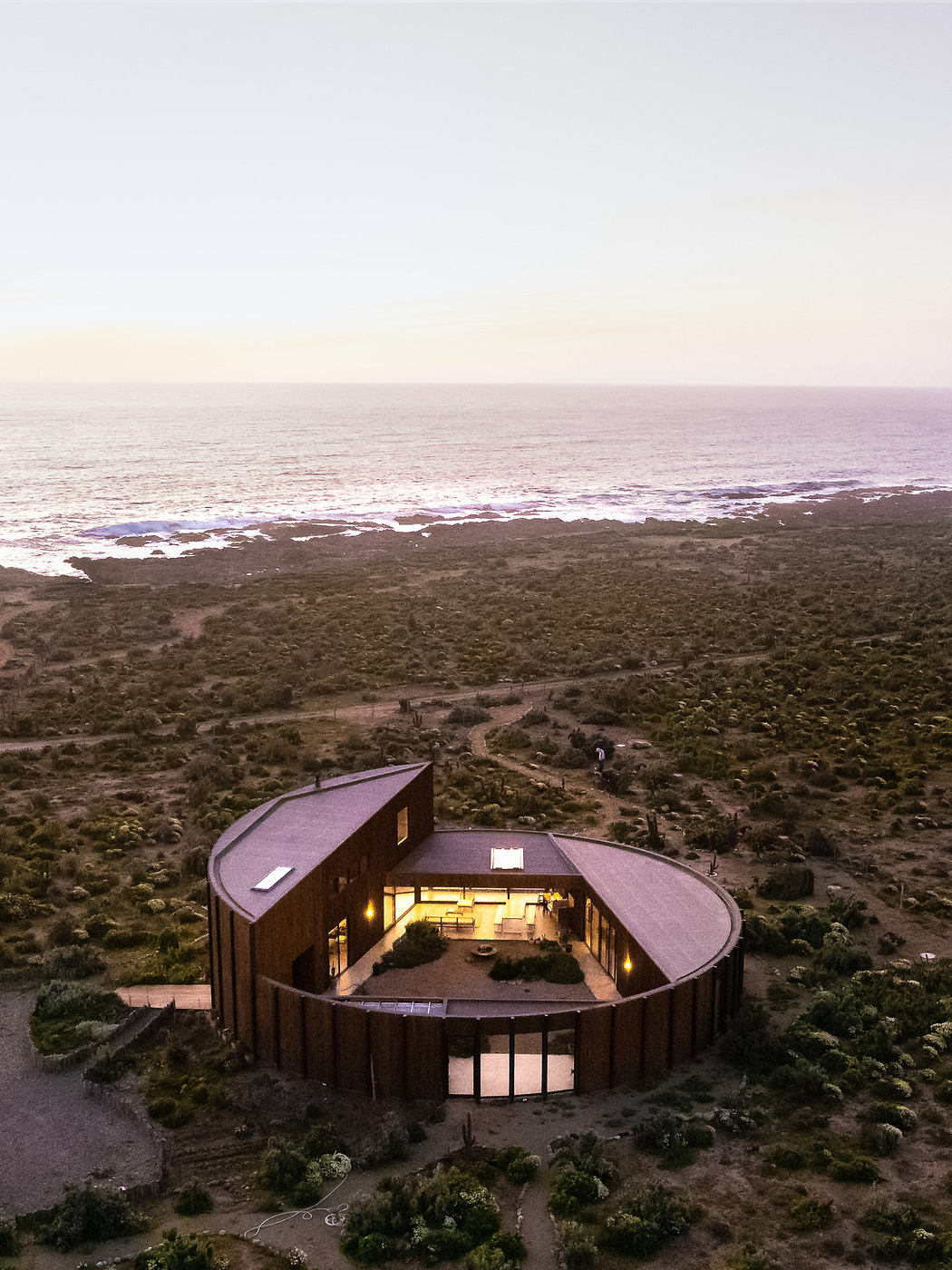 Circular modern home with lit interior by the ocean at dusk.