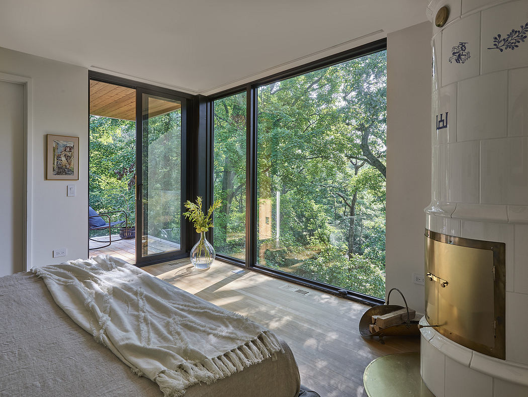 Modern bedroom with large windows overlooking trees and a white tile fireplace.
