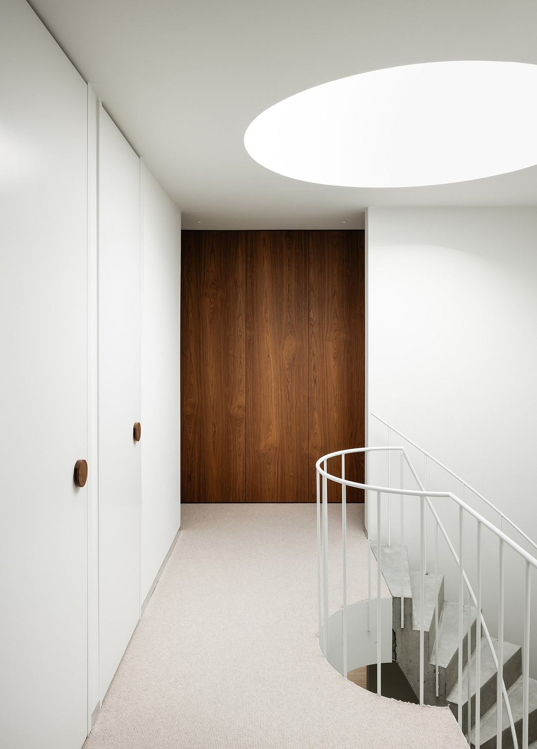 Minimalist hallway with a wooden door, white walls, and circular skylight