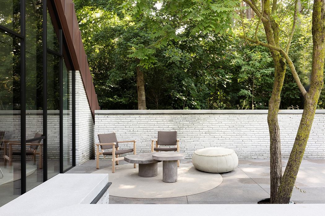 Modern outdoor patio with concrete furniture and large glass windows surrounded by trees.