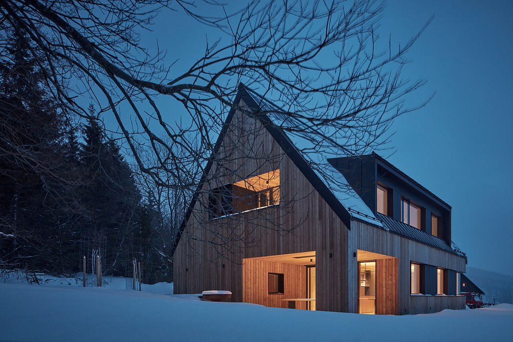Modern wooden house with illuminated windows against a snowy landscape at dusk.