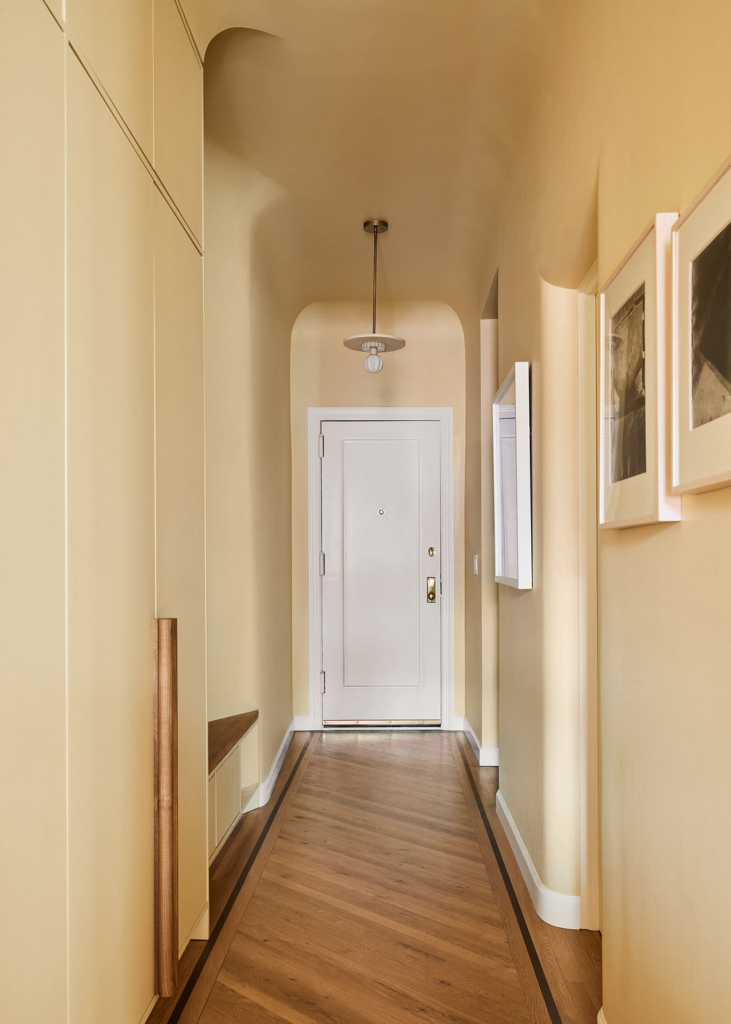 Narrow hallway with wooden floor leading to a white door, framed pictures on wall