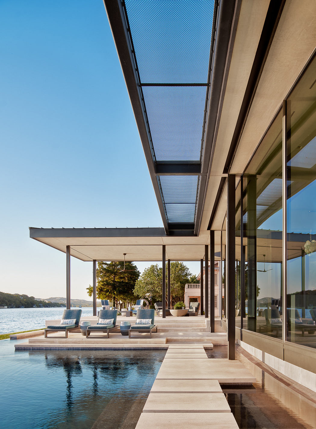 Modern waterfront home with glass walls and outdoor seating area under a pergola.