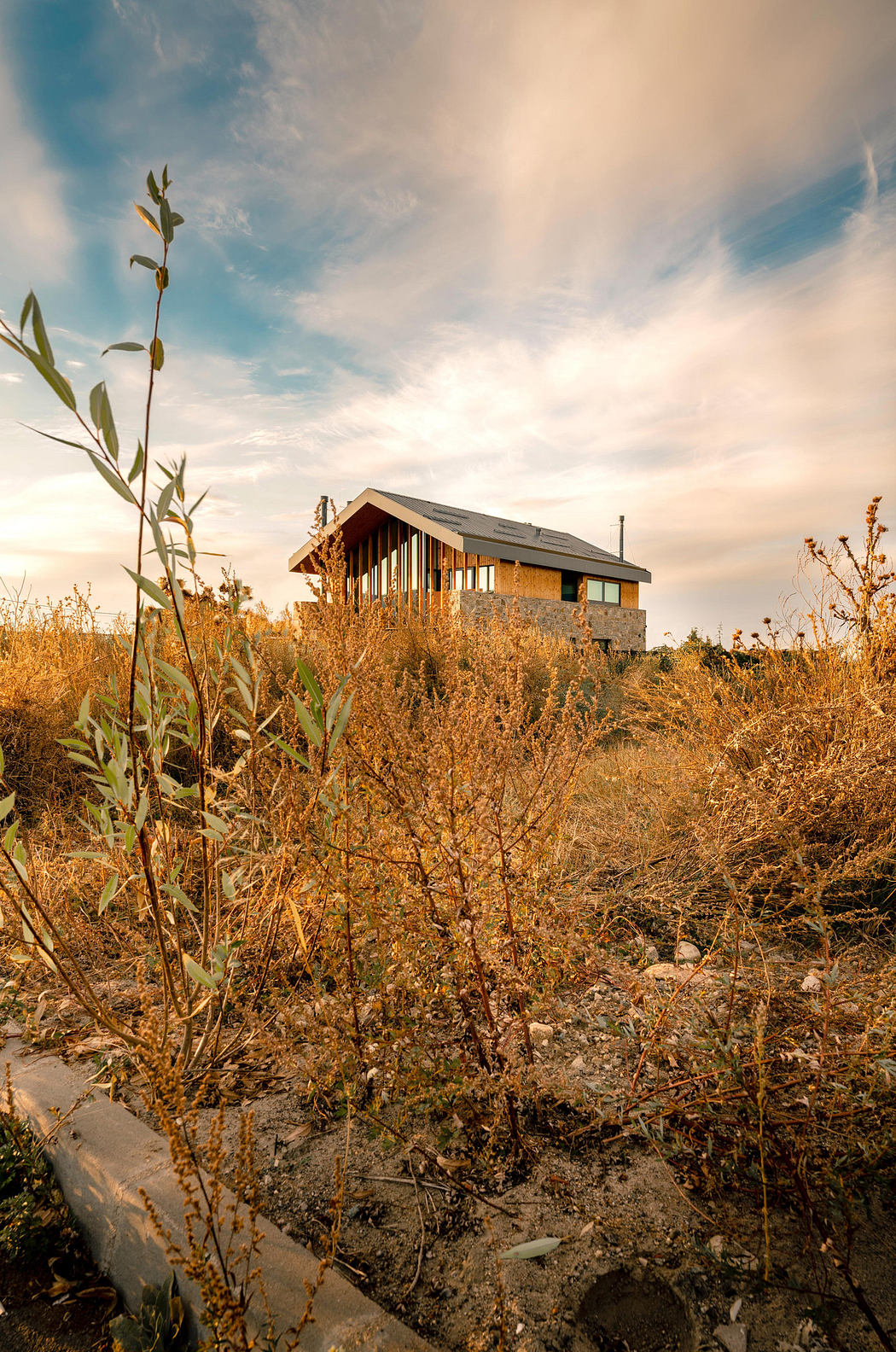 Modern house on a hillside with dry grass and a sunset sky.