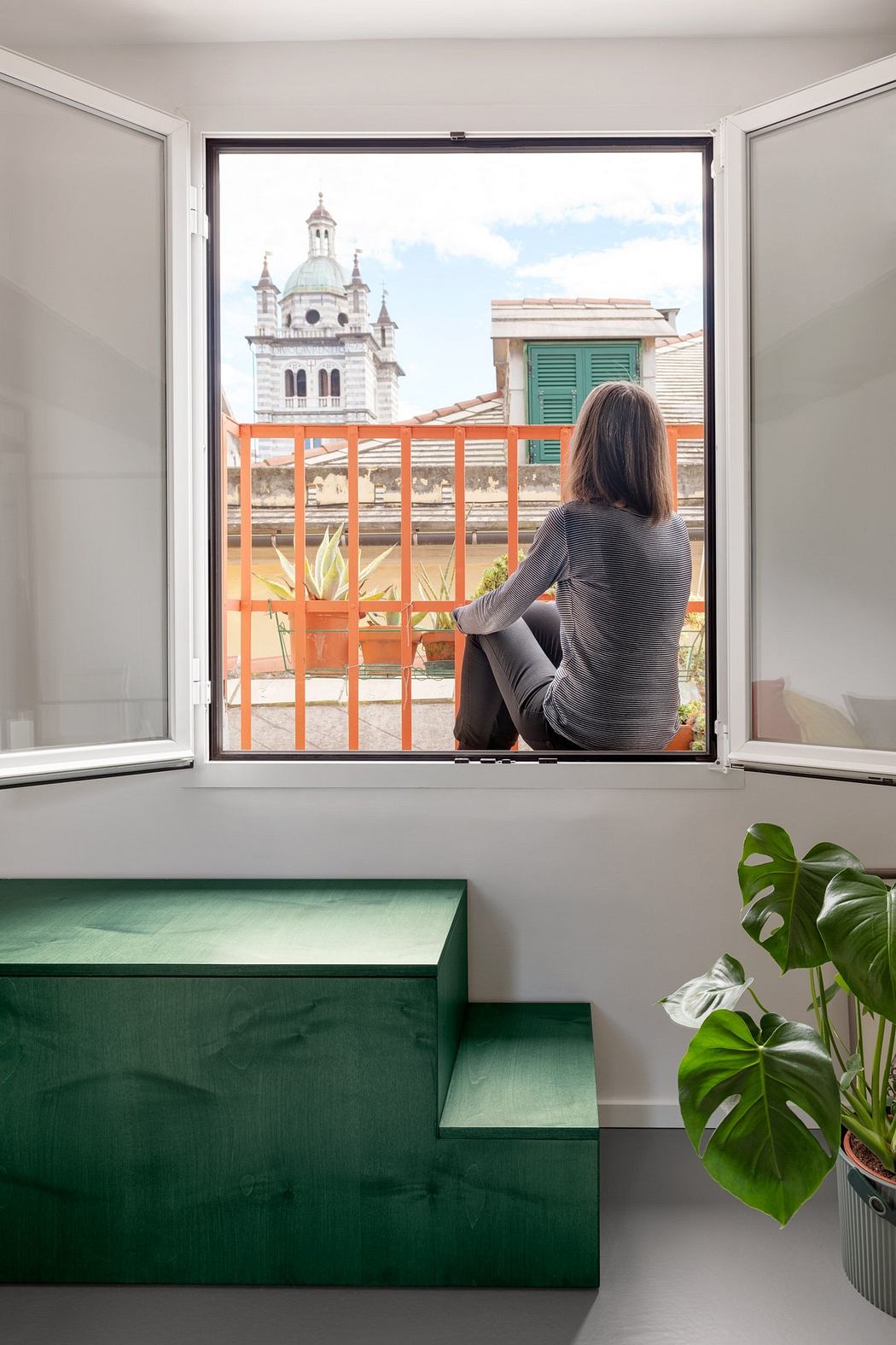 Person seated at a window nook with green steps, overlooking urban scenery.