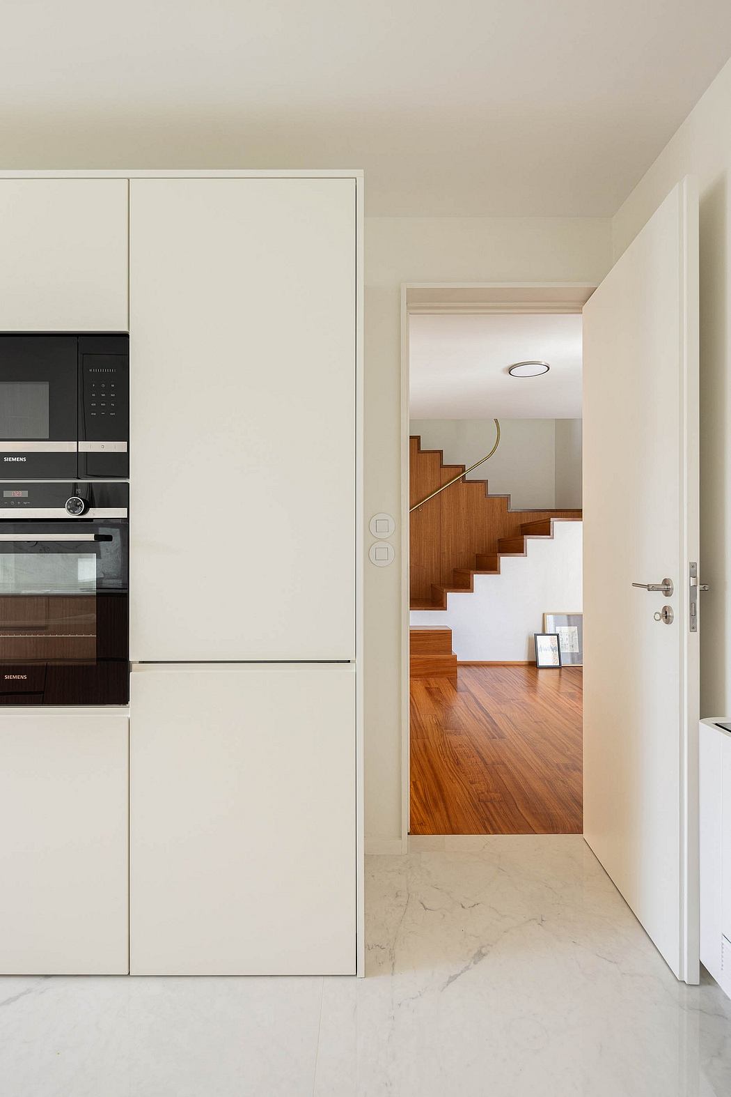 Modern kitchen with built-in appliances and a view into a hallway with wooden stairs.