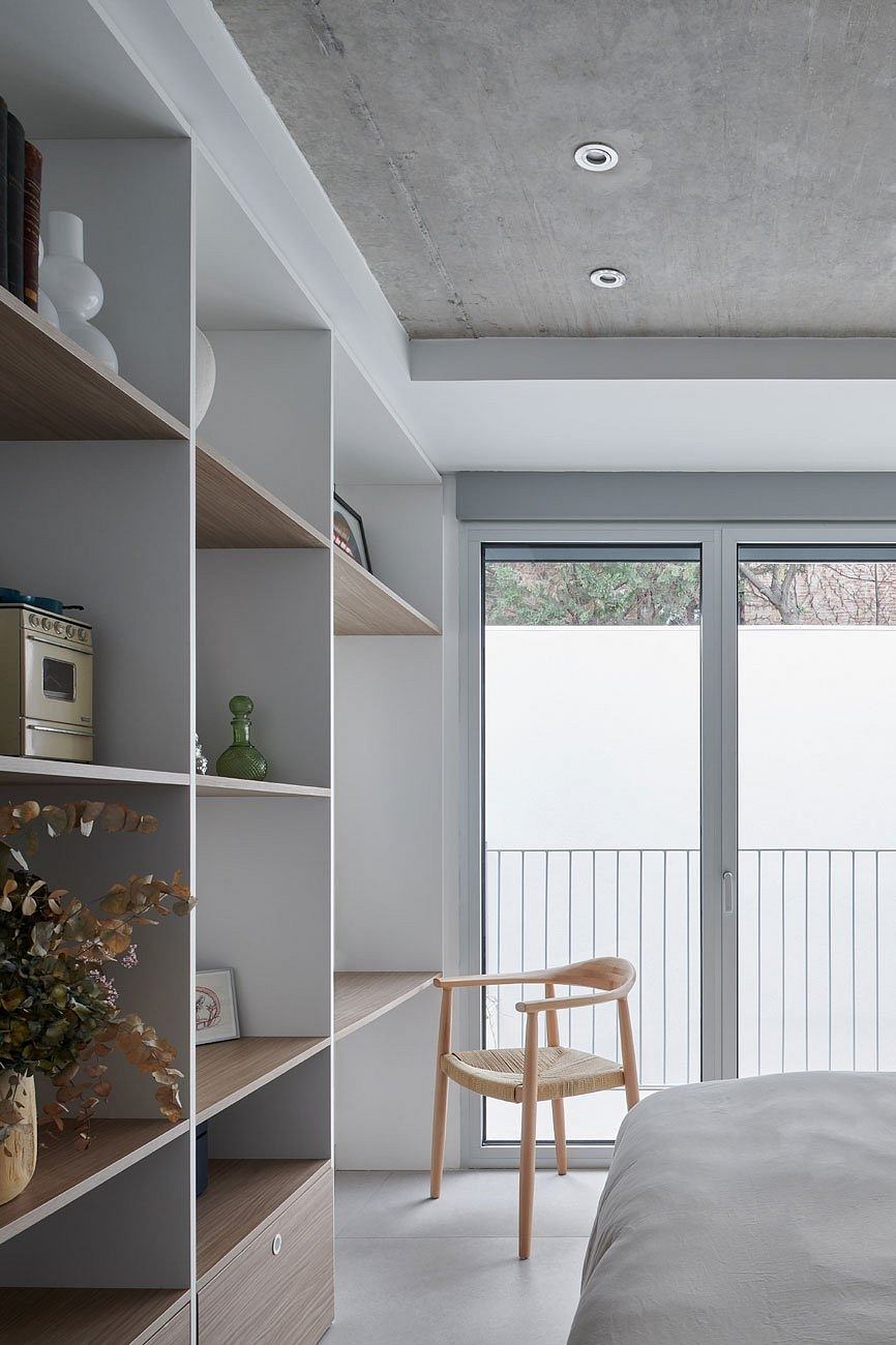 Modern bedroom with white bookshelf, bed, wooden chair and exposed concrete ceiling.