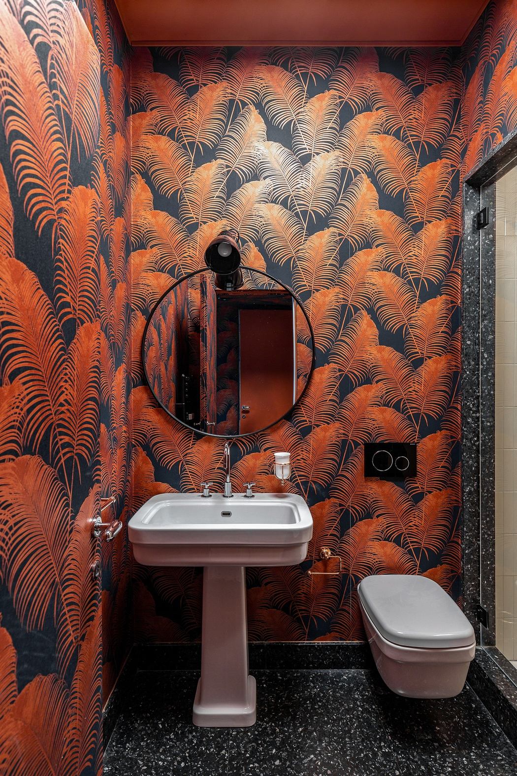 Bathroom with vibrant orange palm wallpaper, pedestal sink, and round mirror.
