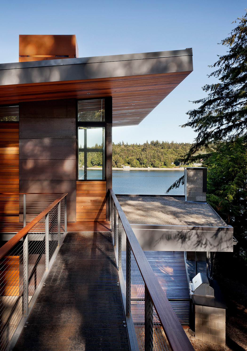 Modern house with wood siding, metal roof, and walkway overlooking a lake.