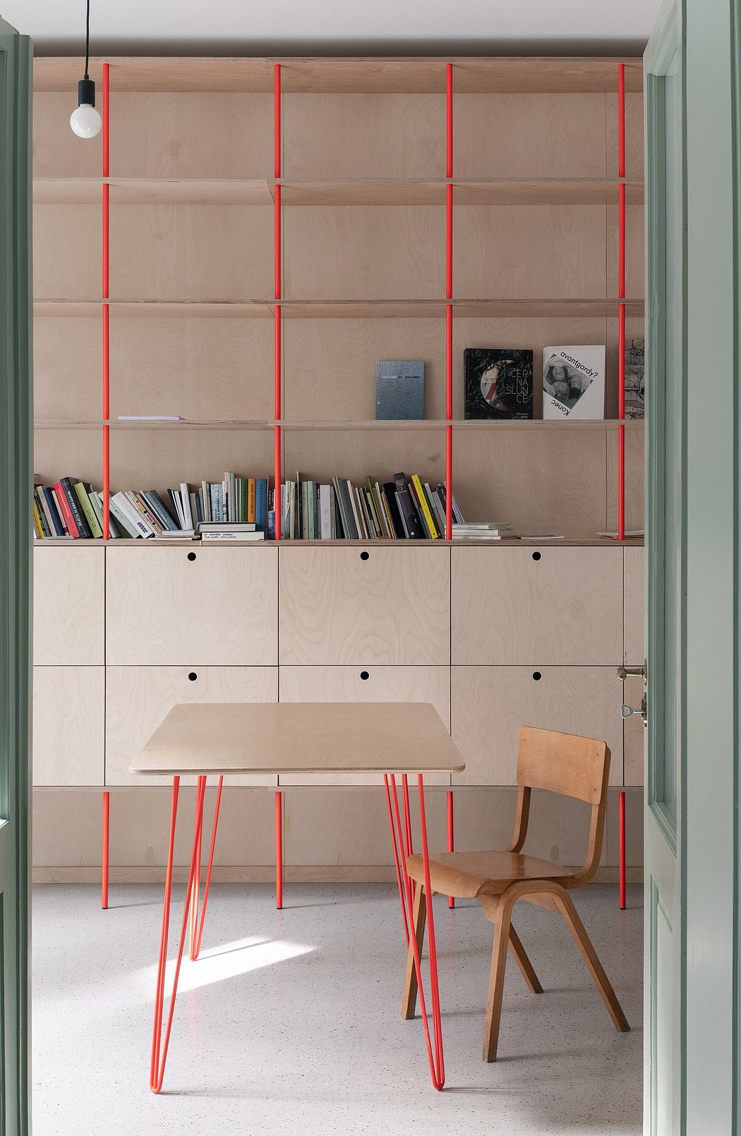 Modern home office with a wooden desk, red legs, matching shelf, and a