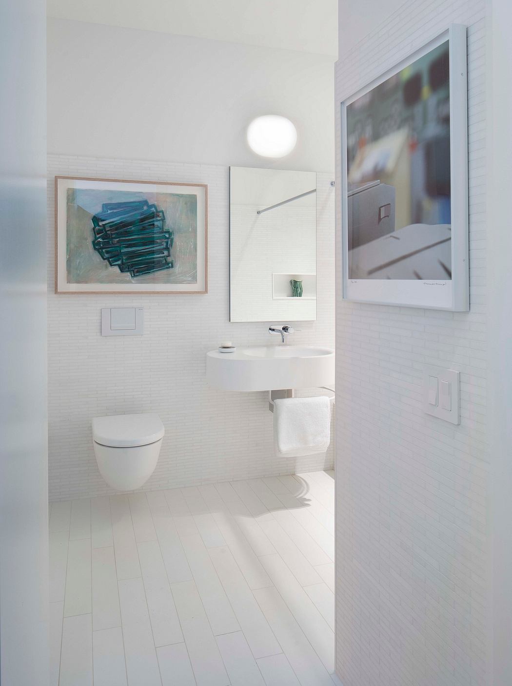 Modern white bathroom with wall-mounted toilet, sink, art, and mirror.