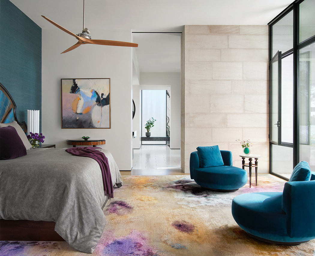 Modern bedroom with blue armchairs, colorful rug, and abstract art.
