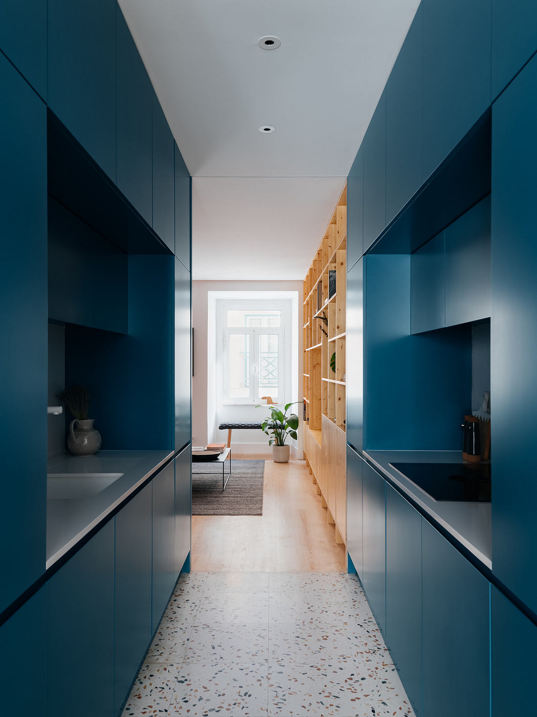 Narrow corridor lined with blue cabinets leading to a bright room with wooden shelving