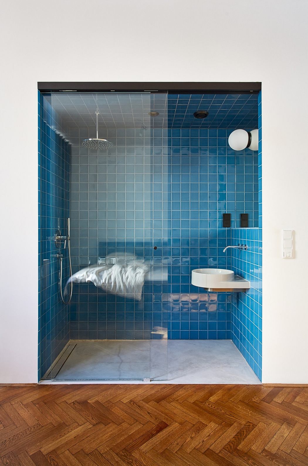 Modern bathroom with blue tiles, glass door, and herringbone wood floor.