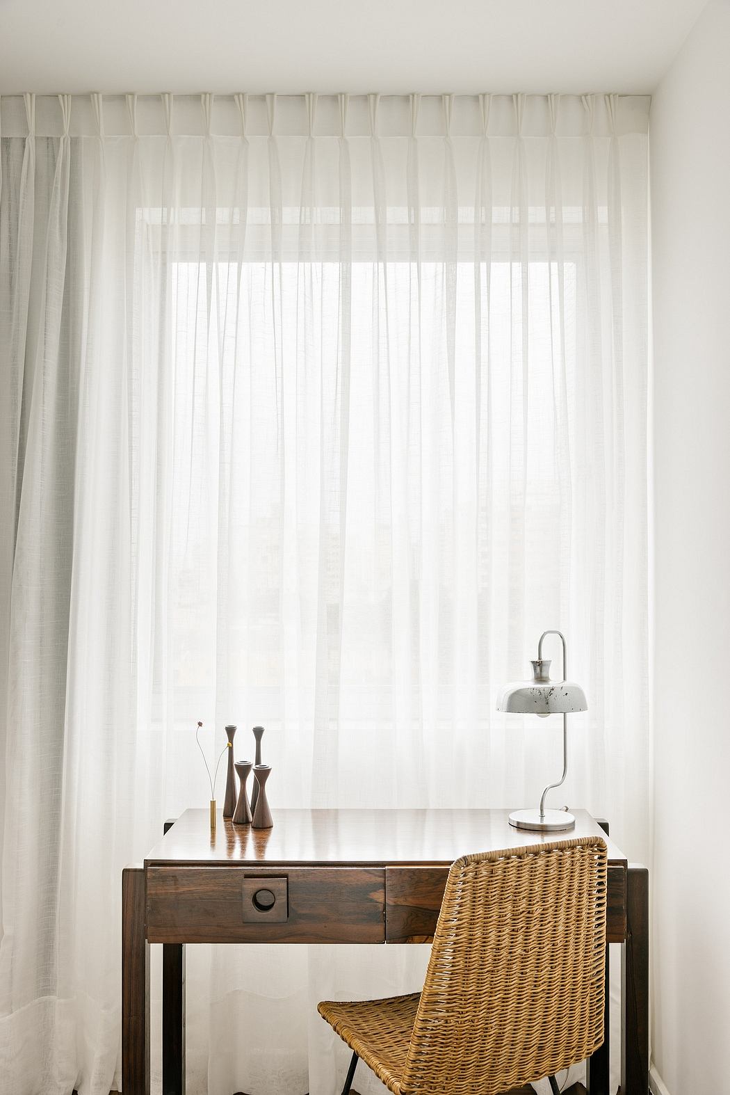 Minimalist study area with wooden desk, wicker chair, and sheer curtains.
