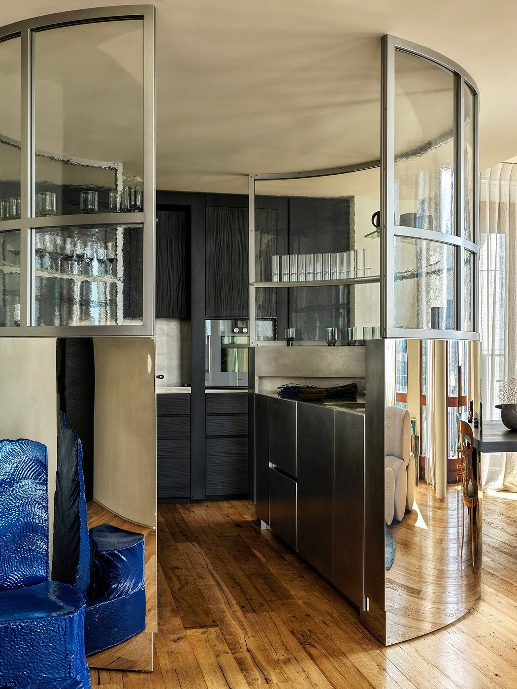 Modern kitchen seen through curved glass doors with wooden flooring.