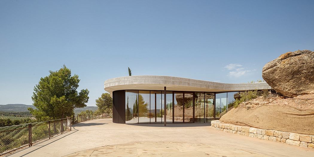 Modern circular pavilion with glass walls next to a giant boulder under clear skies