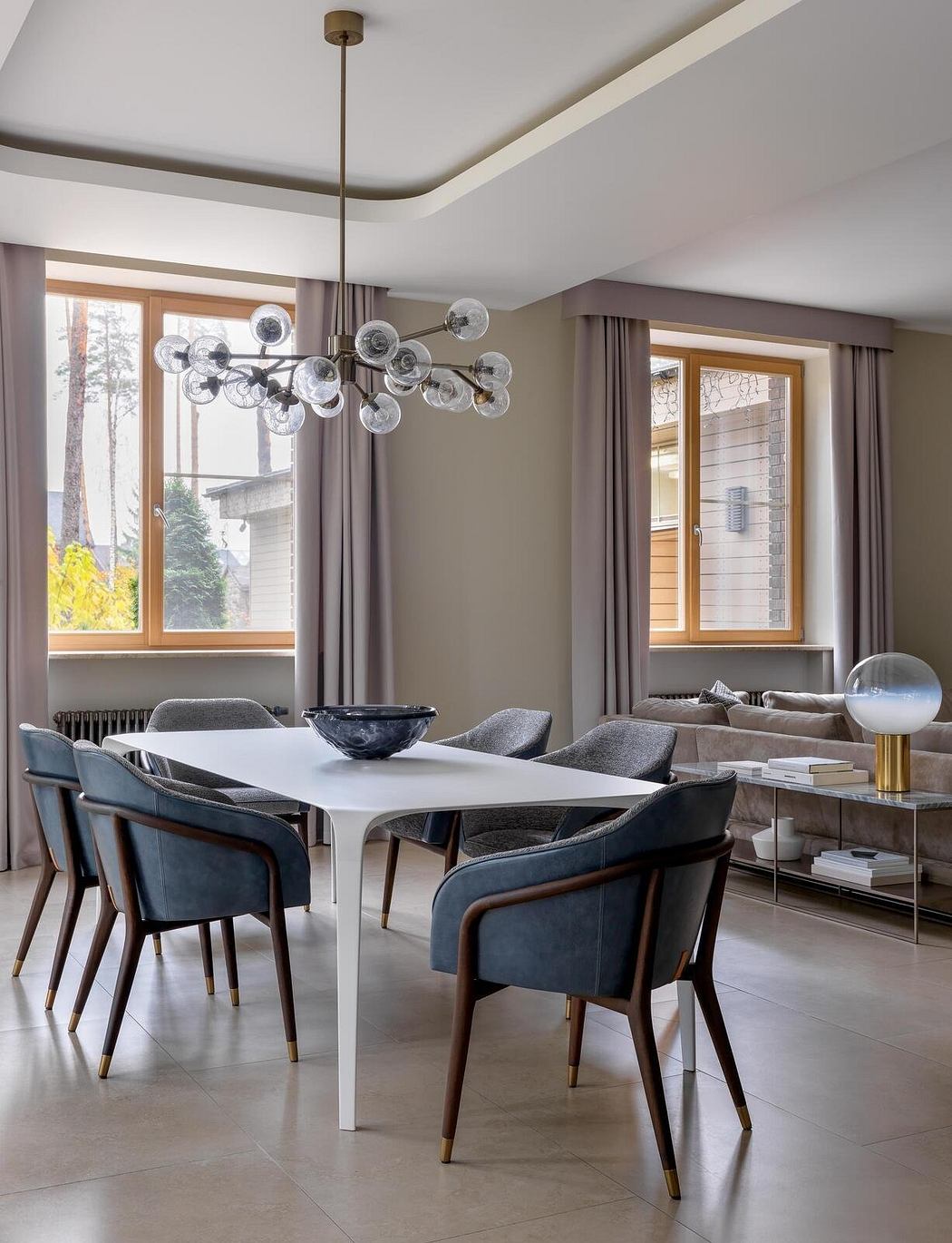 A modern dining area with a large white table, plush grey chairs, and a striking glass chandelier.