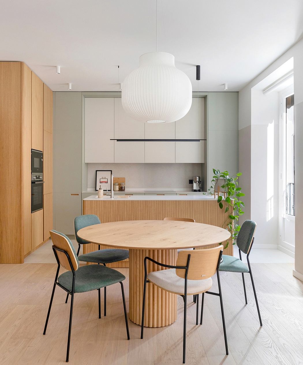 A modern, open-concept kitchen and dining area with a round wooden table, upholstered chairs, and a sculptural pendant lamp.