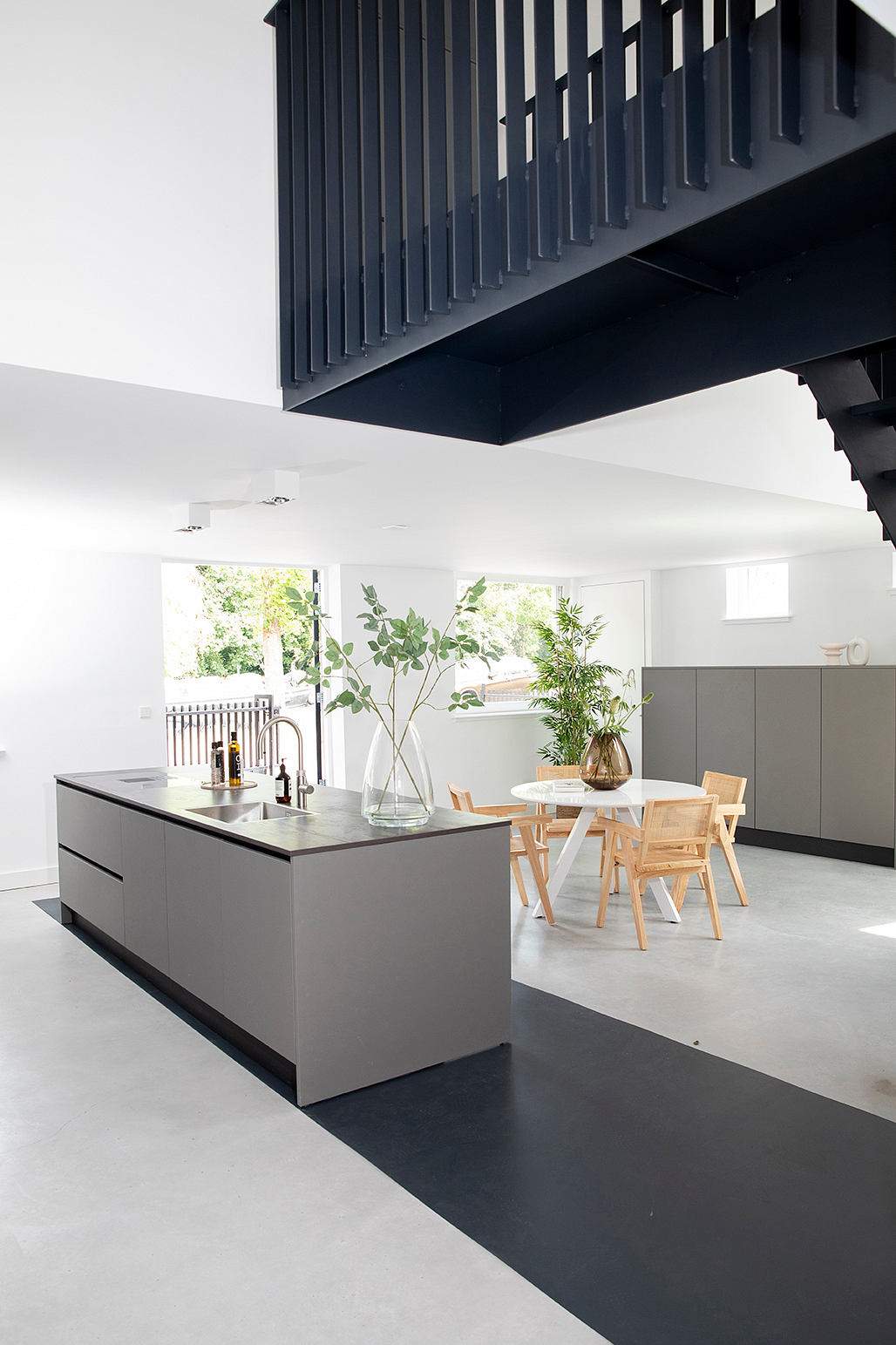 A modern kitchen interior with a sleek grey island, wood dining set, and lush plants.