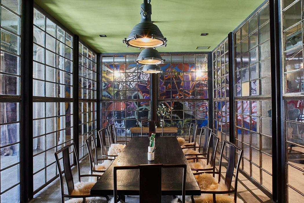 An industrial-style dining room with a long wooden table, pendant lights, and colorful glass panels.