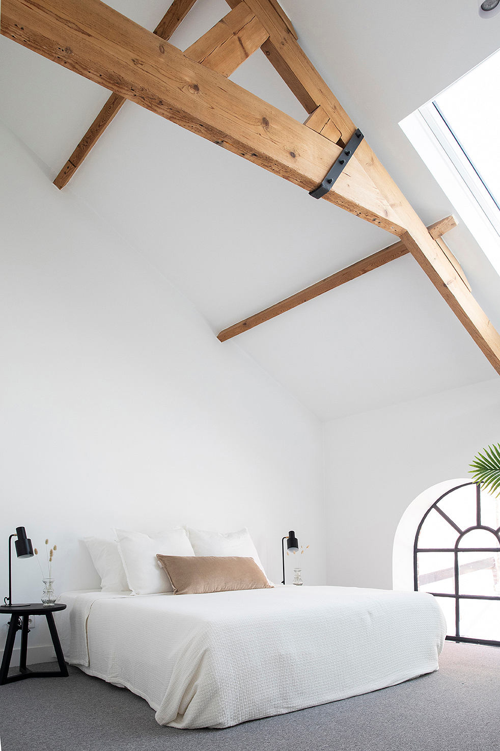 Rustic wooden beams adorn the airy, minimalist bedroom with arched window and neutral tones.
