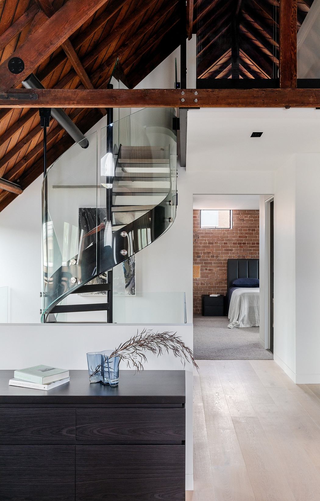 A modern loft with exposed wooden beams, glass staircase, and sleek black furniture.