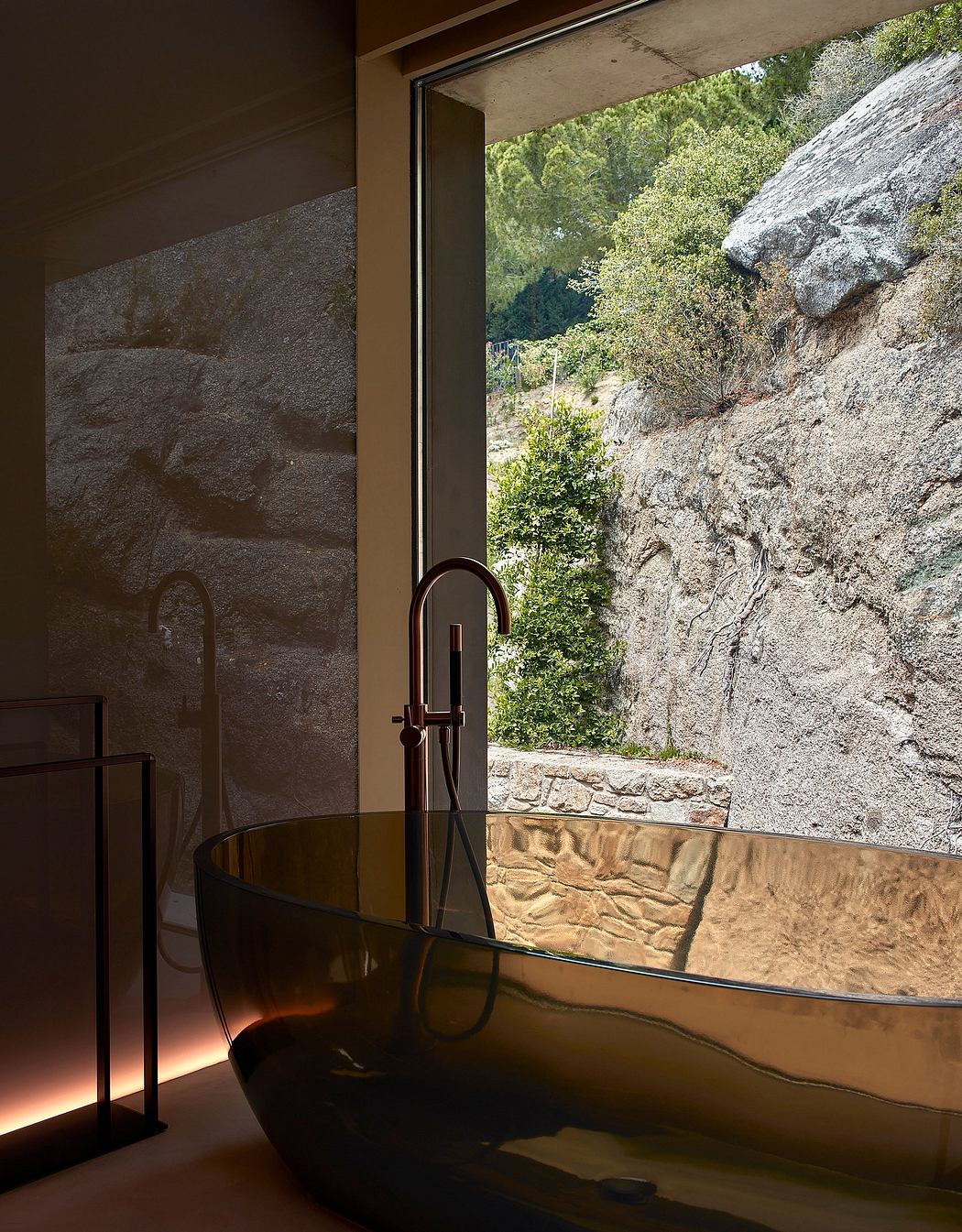 Modern bathroom with a sleek bathtub near a large window overlooking rocks and greenery.