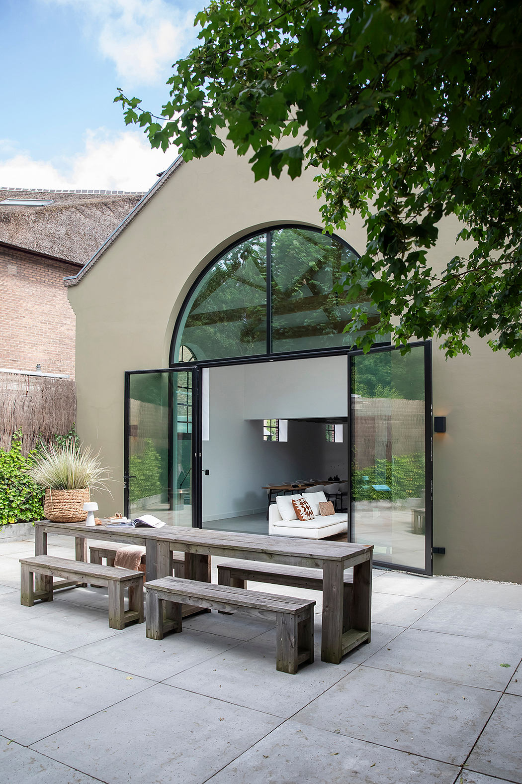 Elegant stone patio with arched glass doors, rustic wooden benches, and lush greenery.