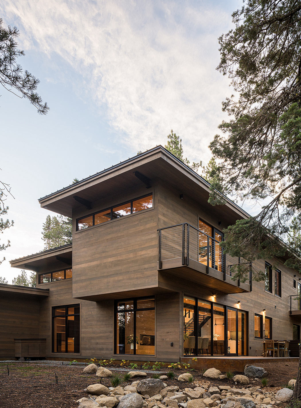 Modern two-story house with large windows and balcony, nestled among trees.