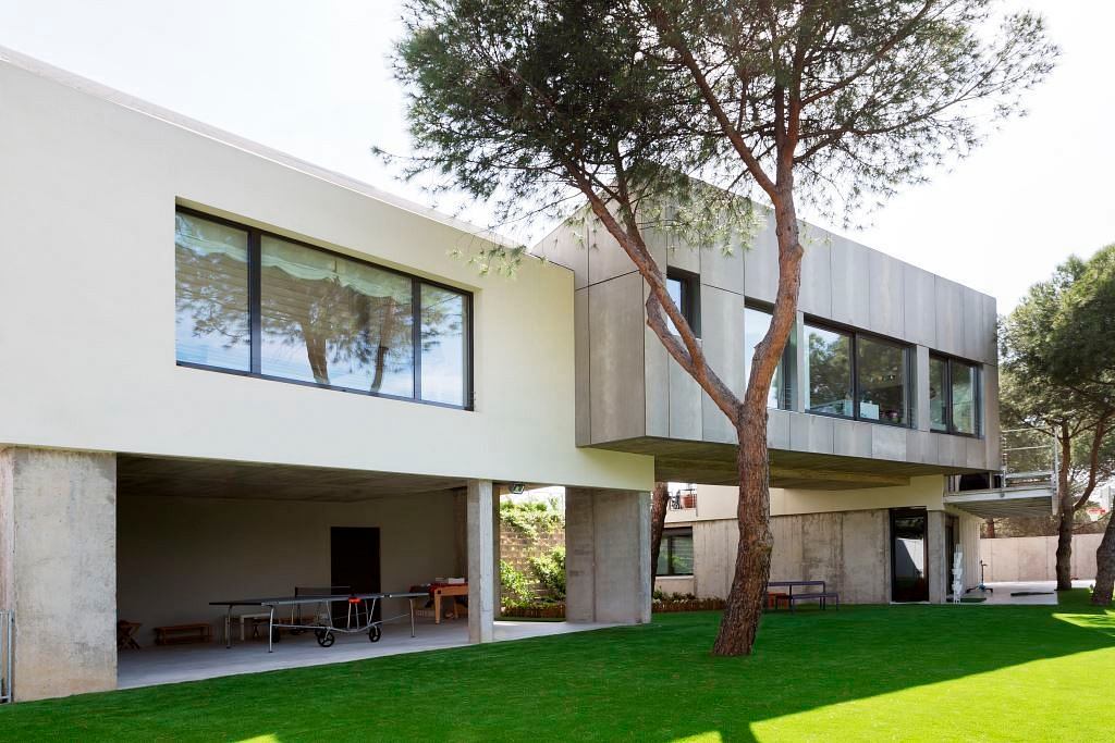Modern two-story house with large windows and a concrete structure, surrounded by green lawn
