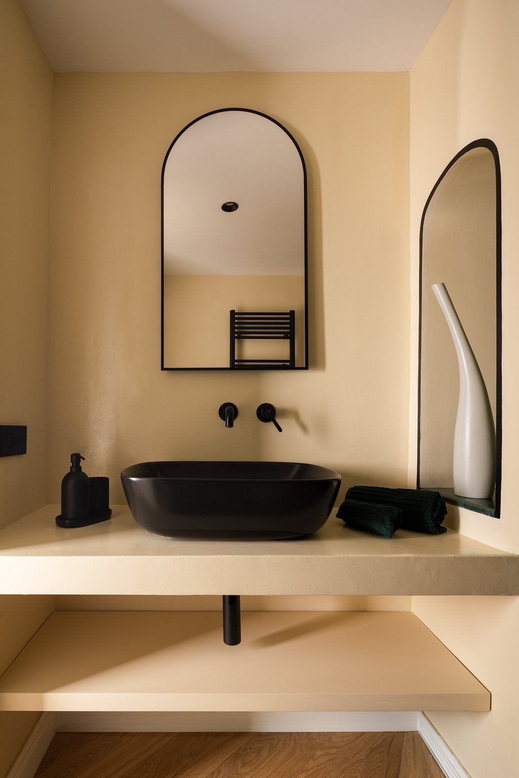 Modern bathroom with beige walls, black basin, and arched mirror.