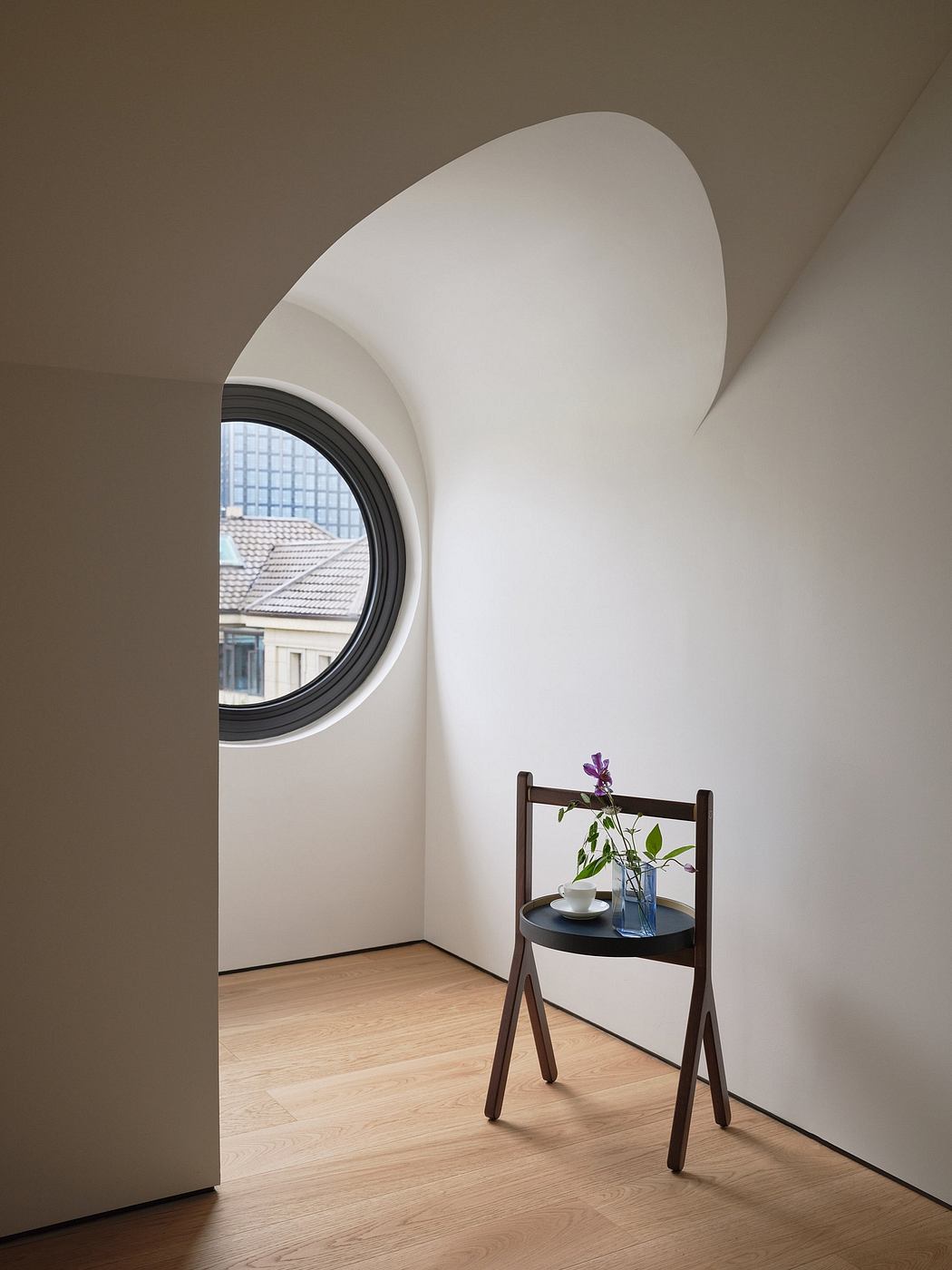 A circular window frames the exterior view, while a minimalist table with a vase of flowers stands below.