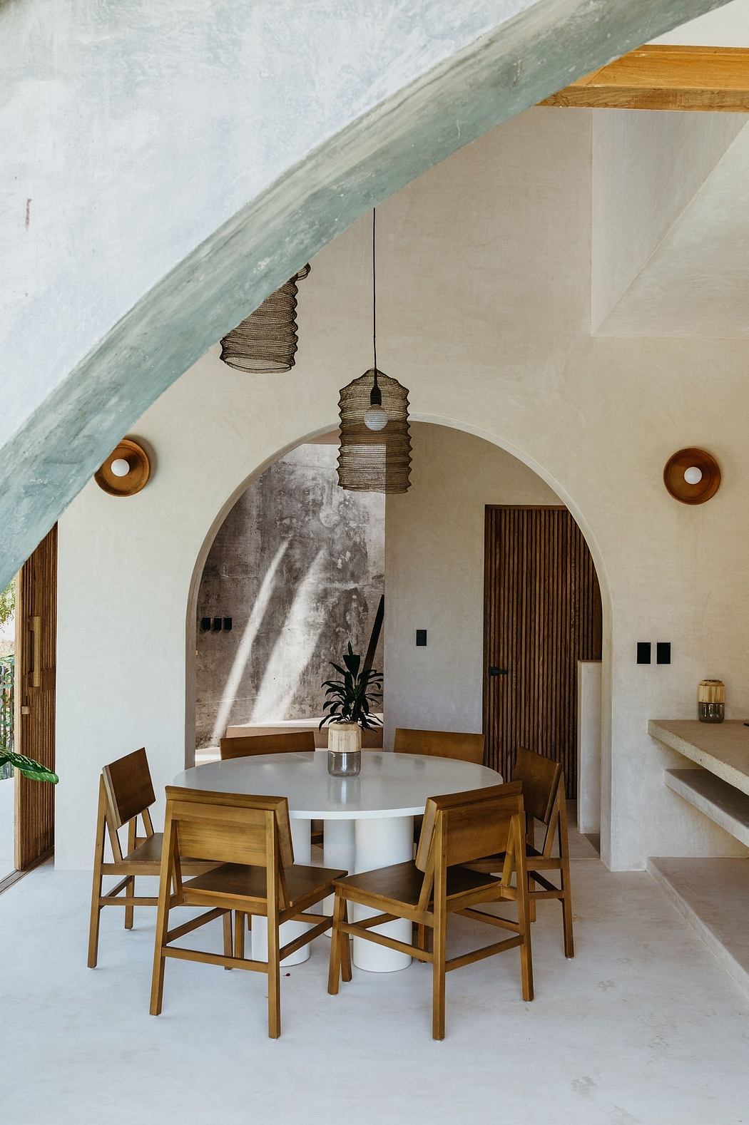 A modern dining area with a round white table, wooden chairs, and pendant lights under a curved ceiling.