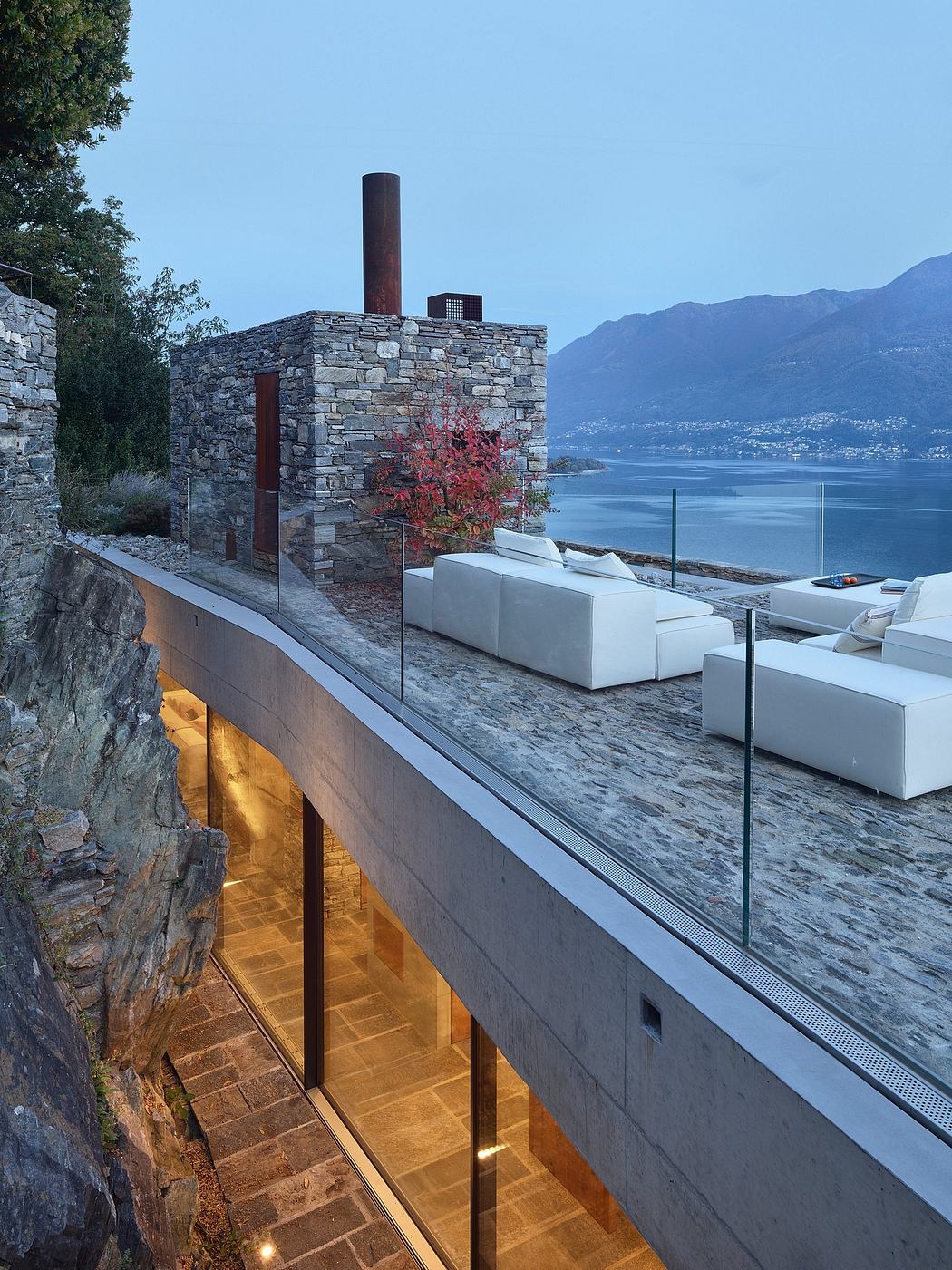 Modern terrace with stone walls, white couches, and lake view at dusk.