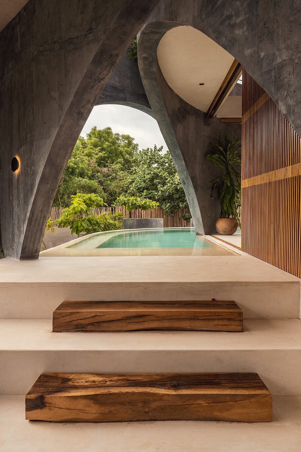 Arched concrete structure frames lush greenery and a serene pool, accented by warm wood elements.