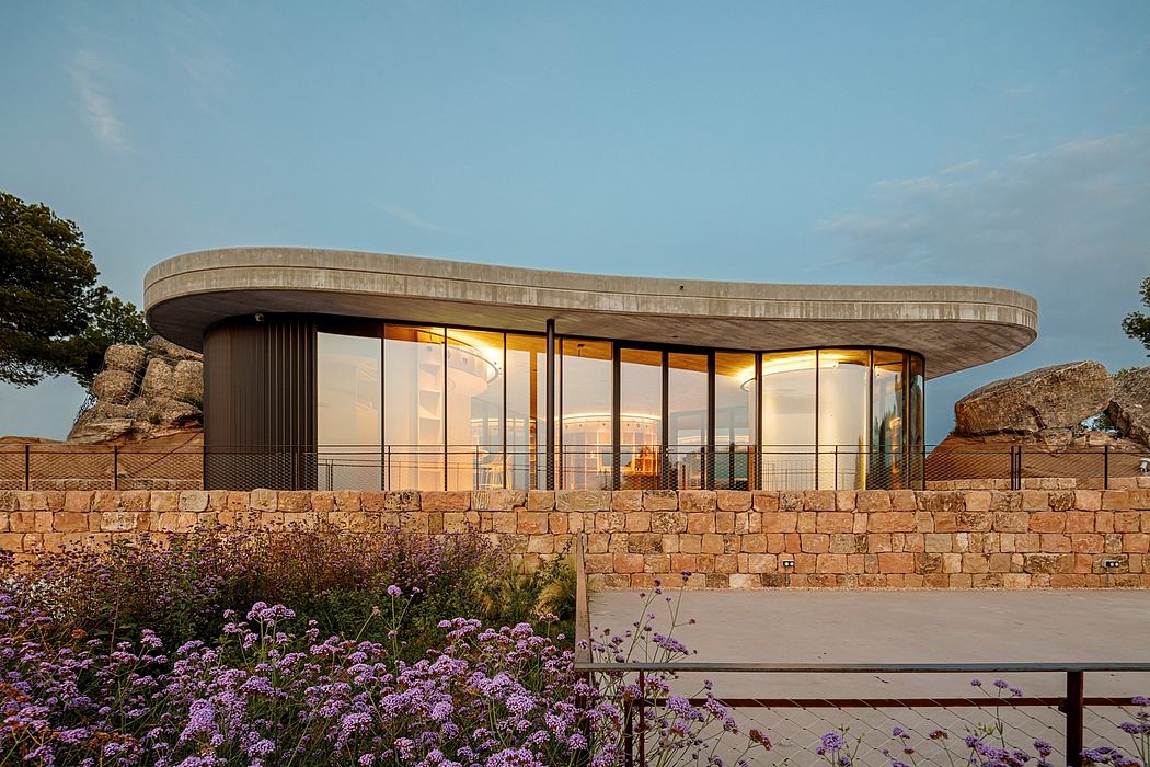 Modern curved house with large glass windows at dusk, surrounded by purple flowers.