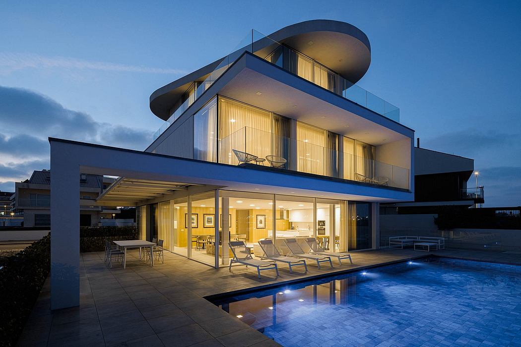 A modern, glass-walled, open-concept home with a pool and lounge chairs at dusk.