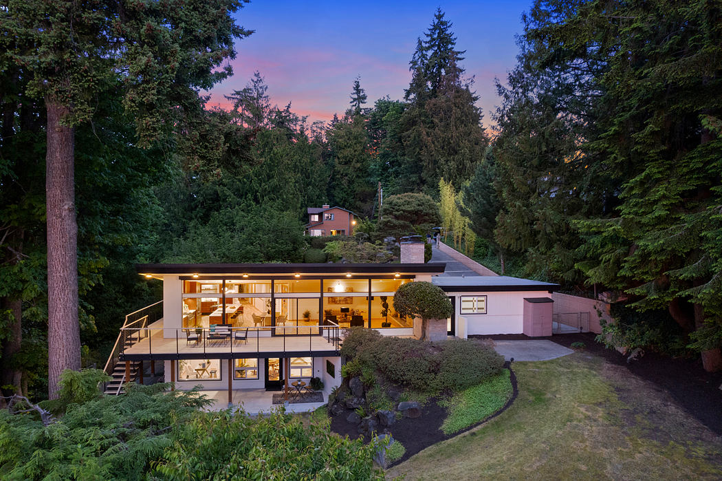 A modern, single-story home with large windows, a deck, and a lush, forested setting.