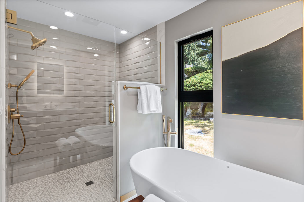 Modern bathroom with sleek tiled walls, large window, freestanding tub, and gold fixtures.